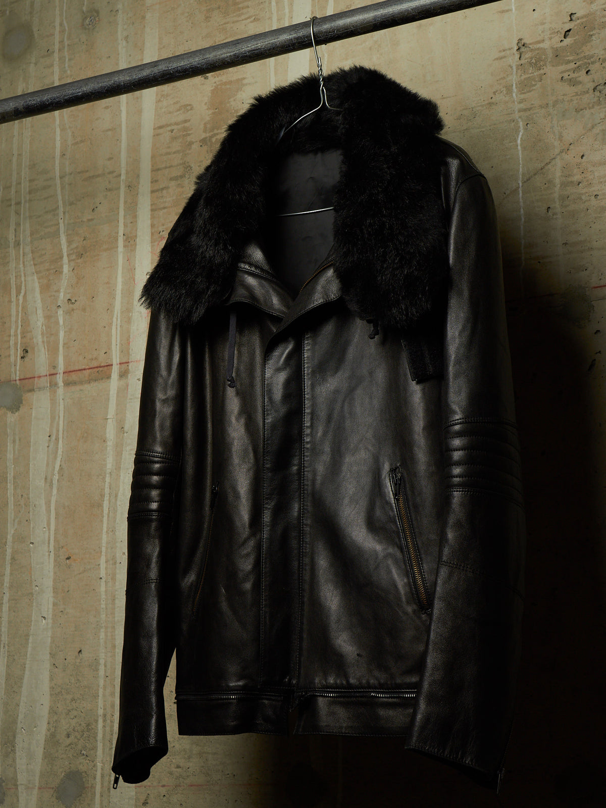 A black leather motorcycle jacket with a thick black faux-fur collar, hanging on a metal hanger from a horizontal bar against a textured concrete wall.