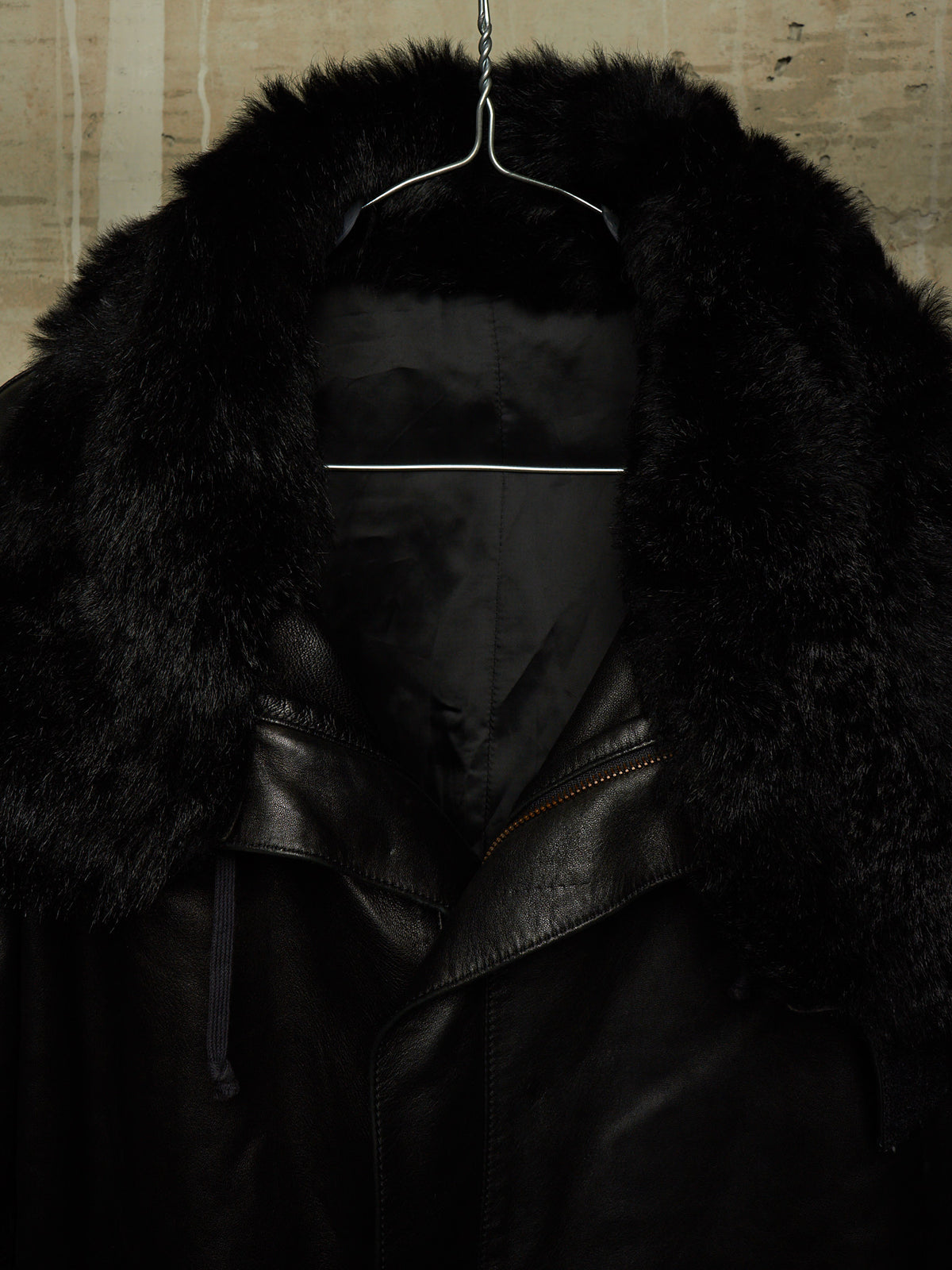 A black leather jacket with a large black fur collar, hanging on a wire hanger against a textured beige wall.