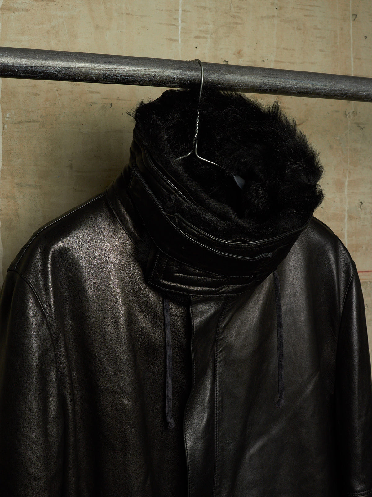A black leather jacket with a high, fluffy black faux-fur collar, hanging on a metal hanger from a horizontal clothing rack against a textured beige wall.