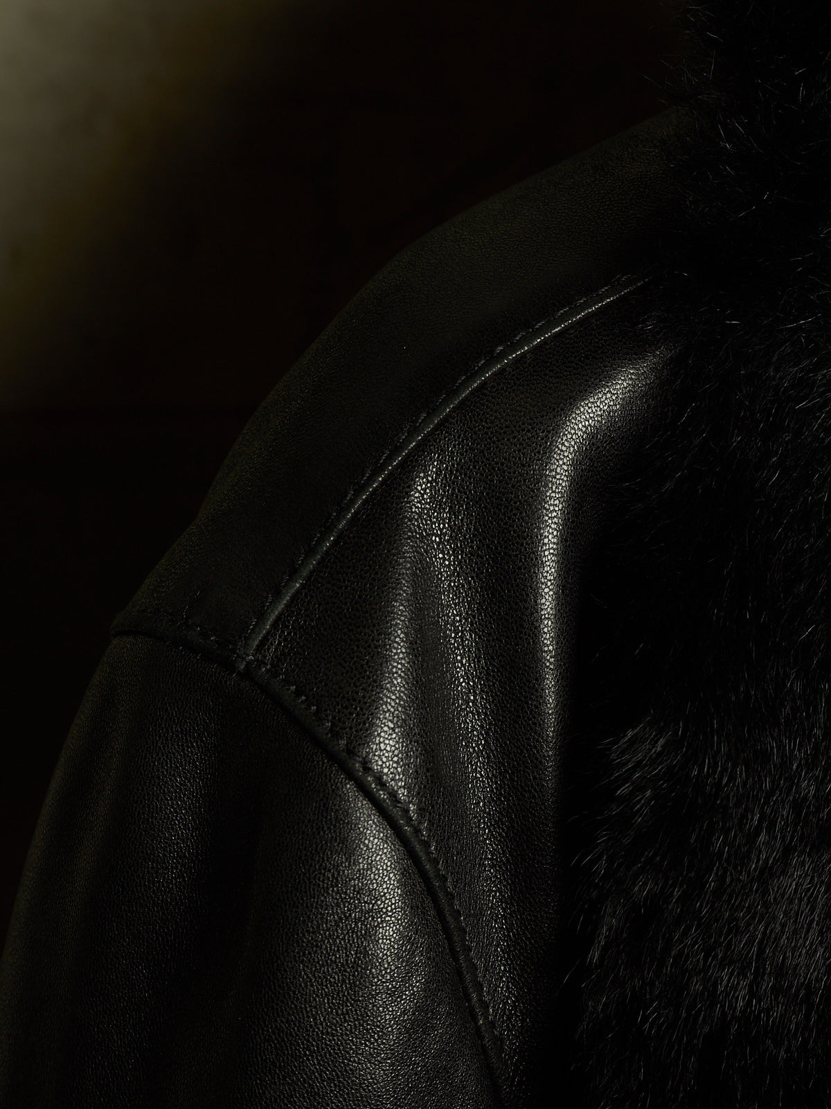 Close-up of a black leather jacket with visible stitching and a dark textured fur or faux-fur panel on the shoulder, photographed in very dark lighting.