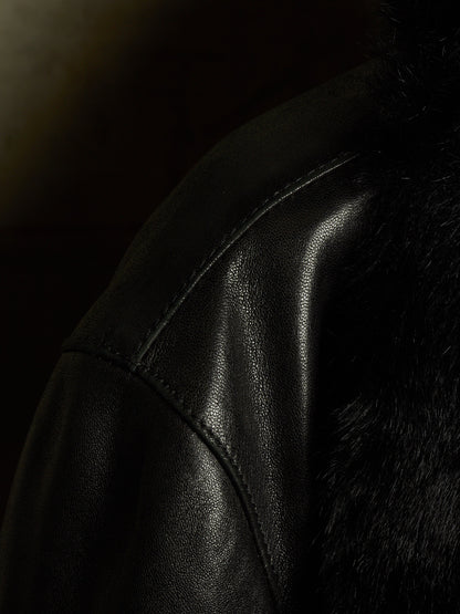 Close-up of a black leather jacket with visible stitching and a dark textured fur or faux-fur panel on the shoulder, photographed in very dark lighting.