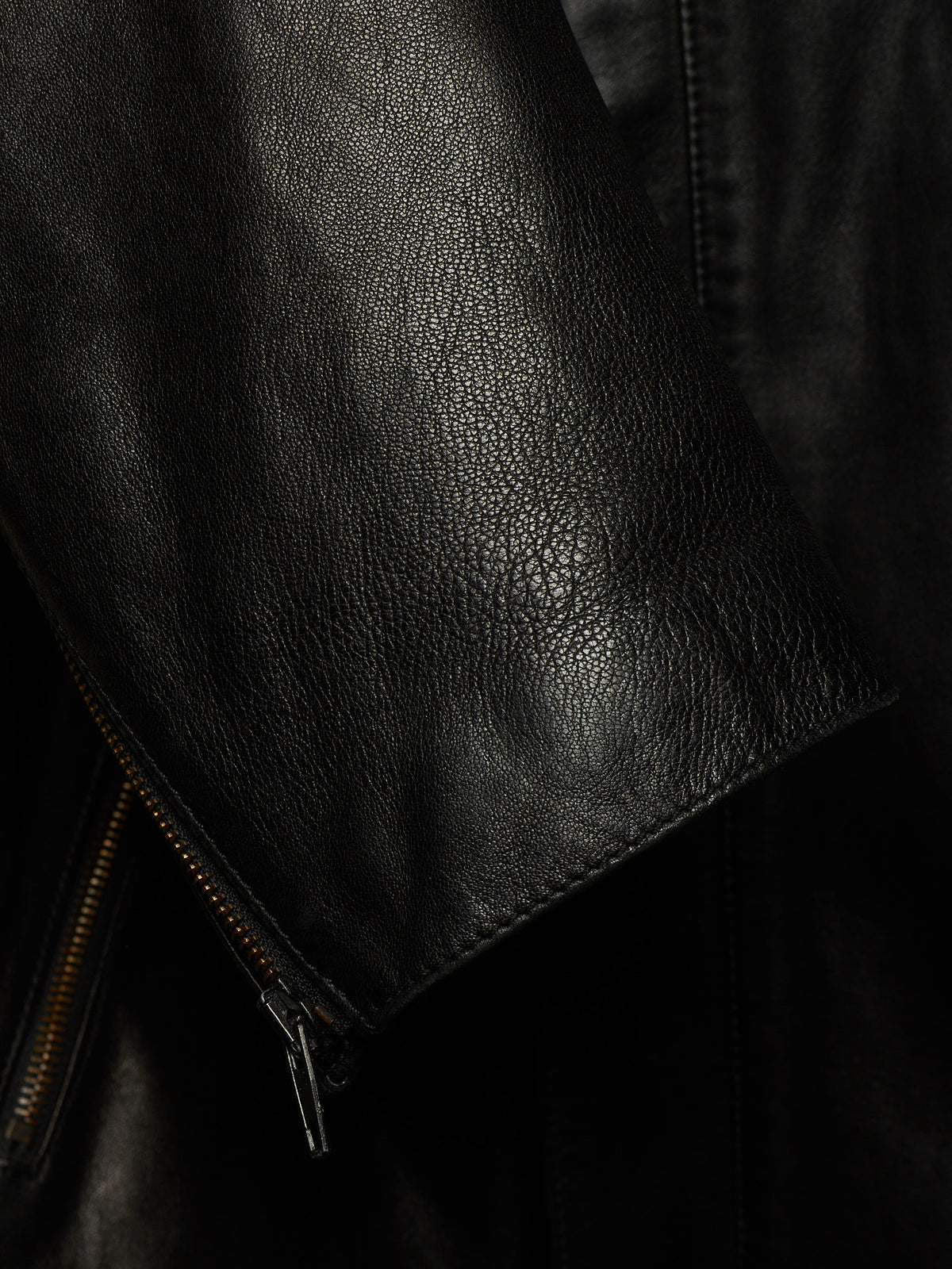 Close-up of a black leather jacket showing the pebbled texture, a zippered sleeve cuff, and dark background.
