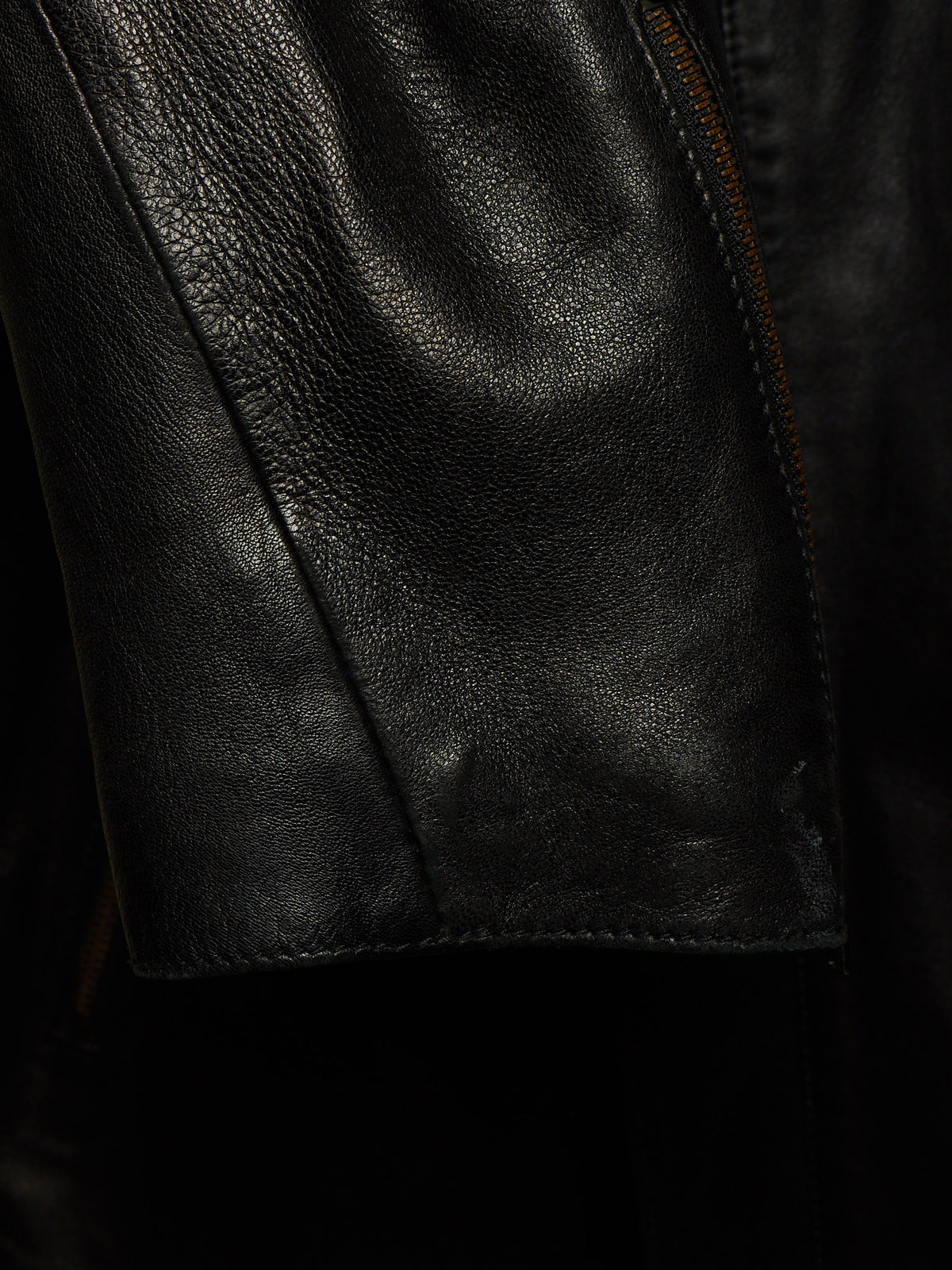 Close-up of a black leather jacket showing the textured grain, a seam, and a zipper detail.