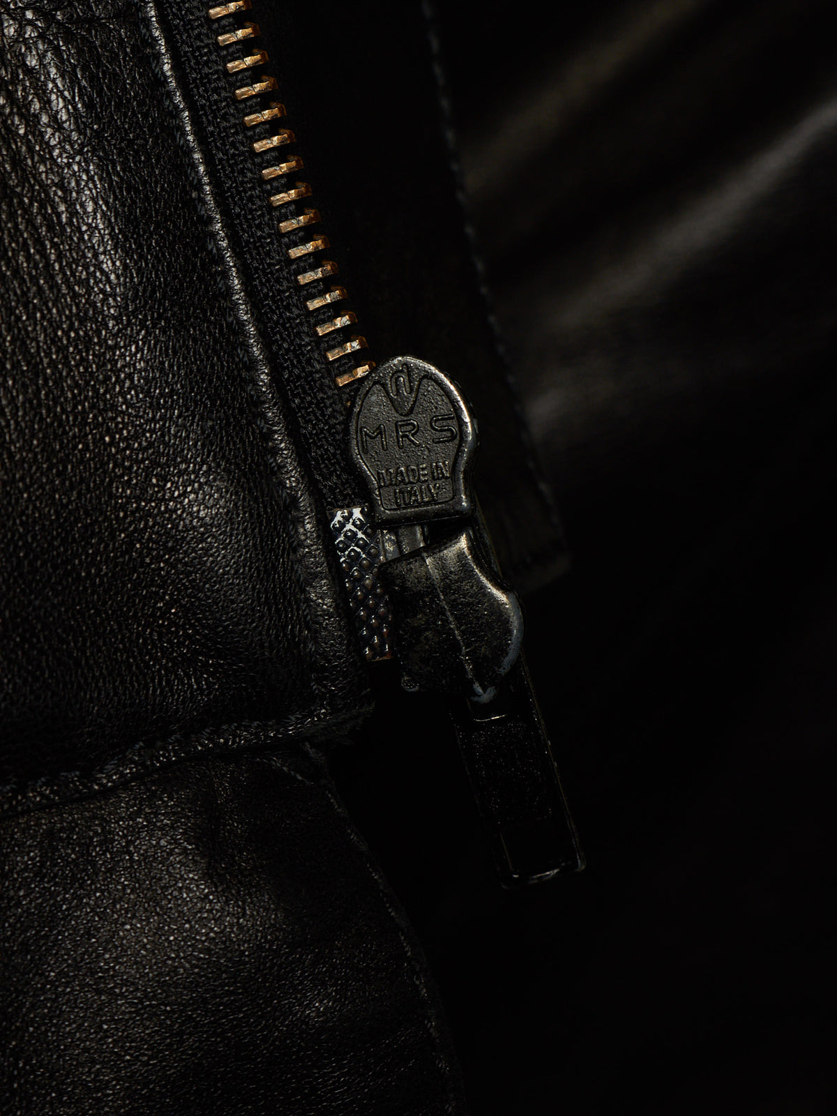 Close-up of a black leather zipper with a dark metal pull tab engraved with 'RS MADE IN ITALY'.