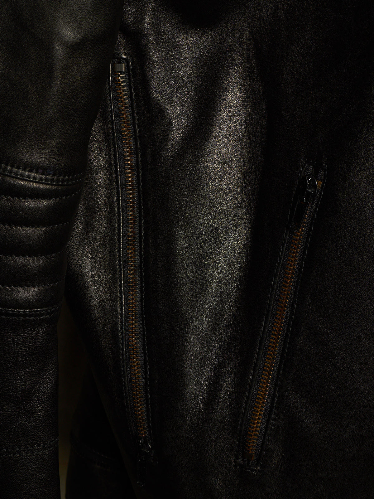 Close-up of a black leather jacket with two vertical zippered pockets and contrast stitching.