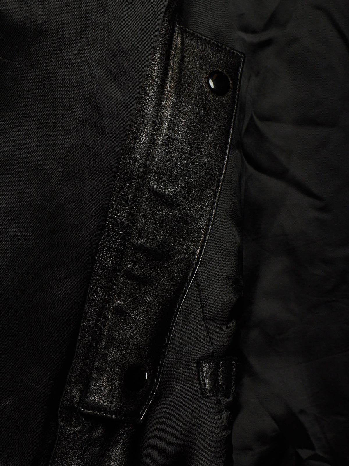 Close-up detail of a black jacket showing a smooth leather-like flap with snap buttons over a matte black fabric body.