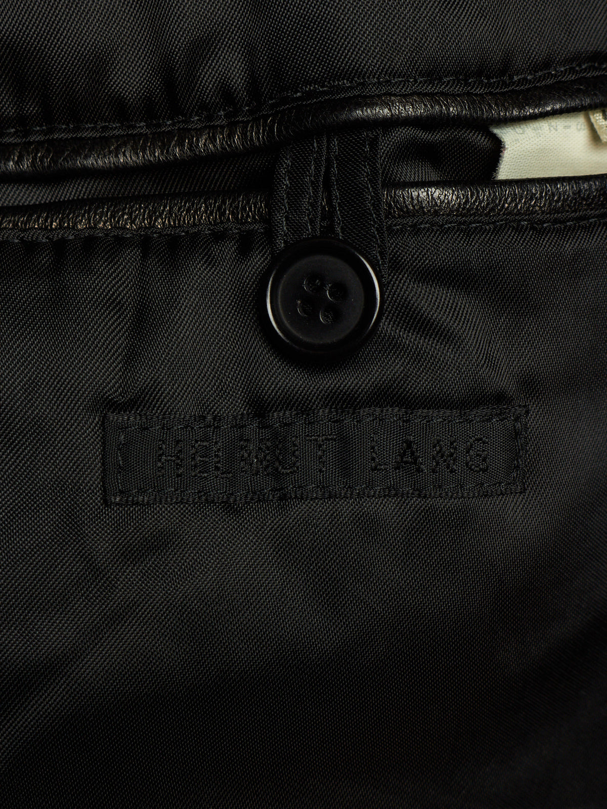 Close-up of a black quilted jacket or puffer garment showing a black button, stitched seam, and a rectangular embroidered label reading HELMET LANG.