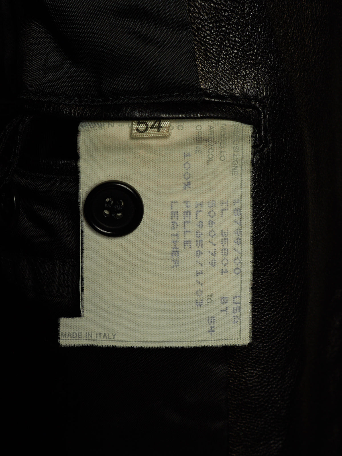 Close-up of a garment interior label sewn onto black leather or faux-leather fabric, with a black button and printed size/manufacturer text visible.