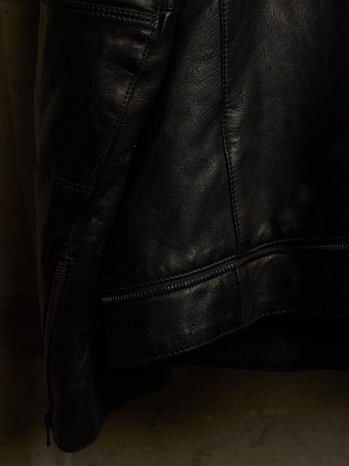 Close-up of a black leather jacket showing the texture, stitching, and a zipper detail against a dark background.