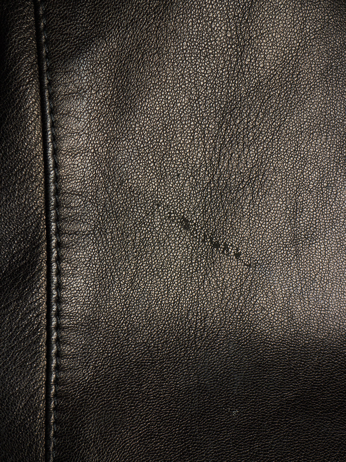 Close-up of black leather texture with visible grain, stitching along the left edge, and a small scratch-like mark across the center.