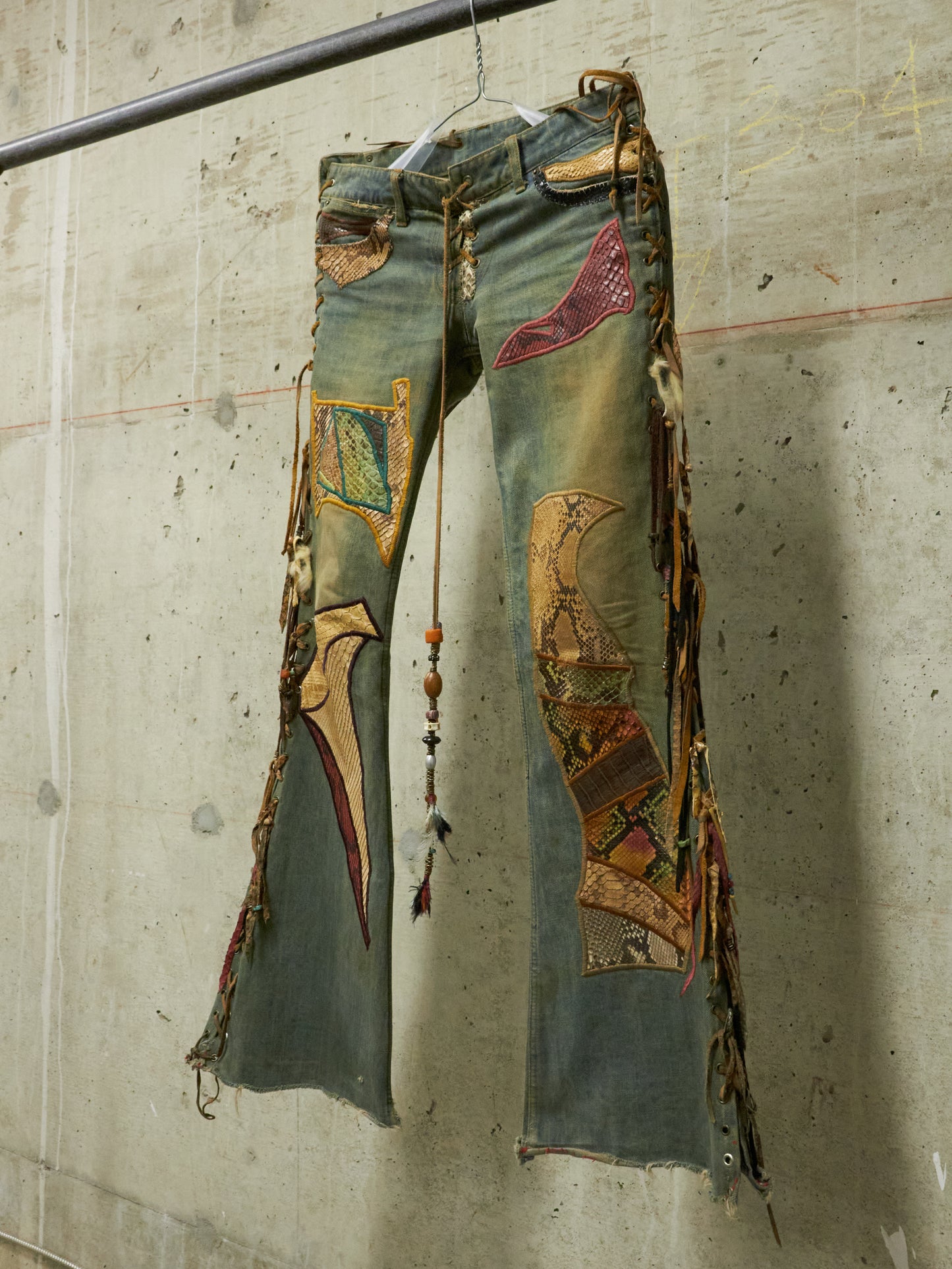 Decorative jeans with patches and fringe hanging against a concrete wall.