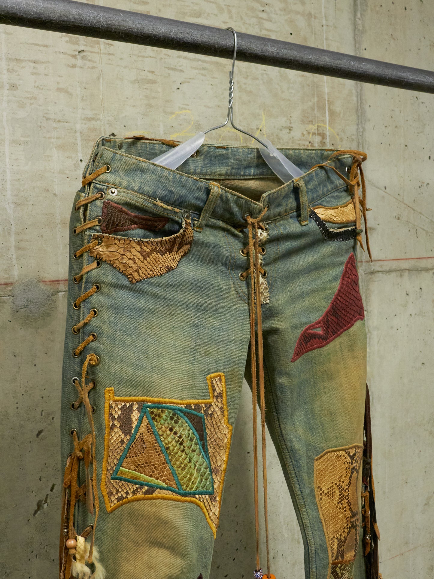 Decorative jeans with patches and embroidery on a hanger against a concrete wall.