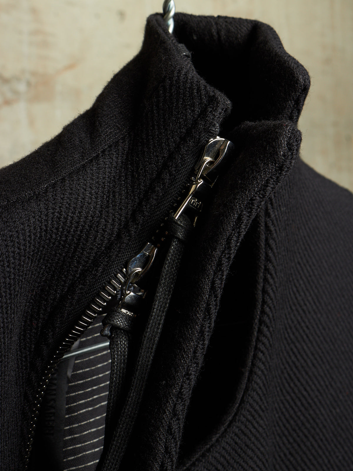 Close-up of a black ribbed knit garment with a silver zipper and metal pull, shown against a light textured background.