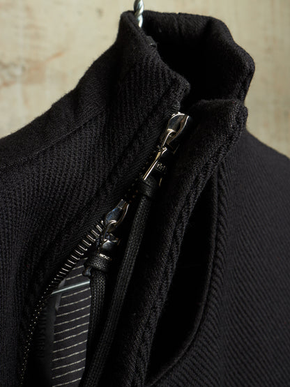 Close-up of a black ribbed knit garment with a silver zipper and metal pull, shown against a light textured background.