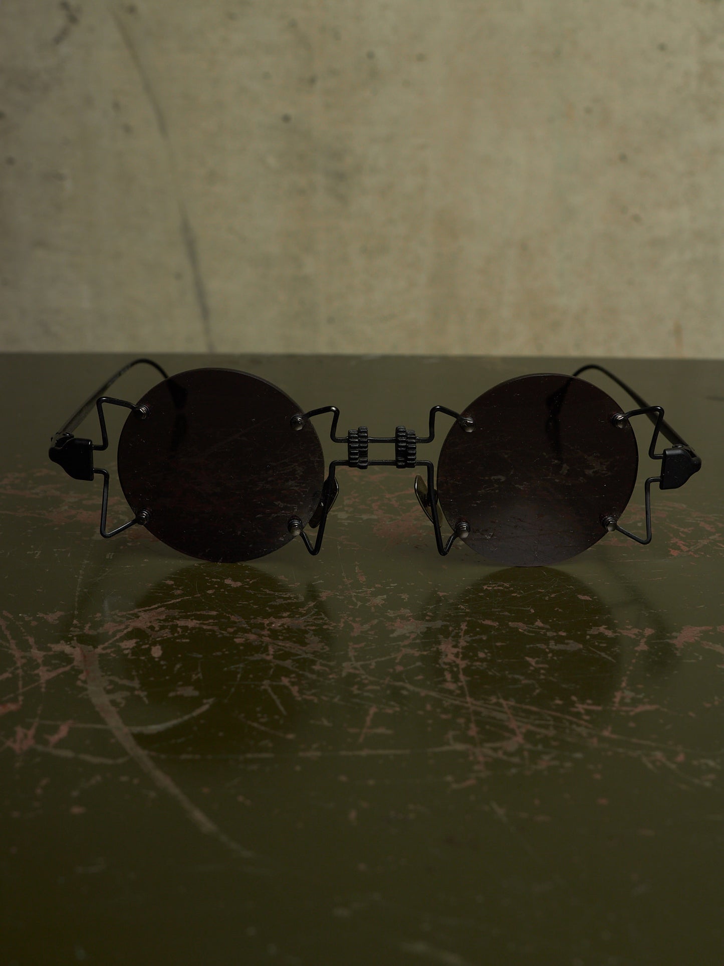 Round black sunglasses with a unique wire frame design, resting on a dark scratched surface against a textured beige background.