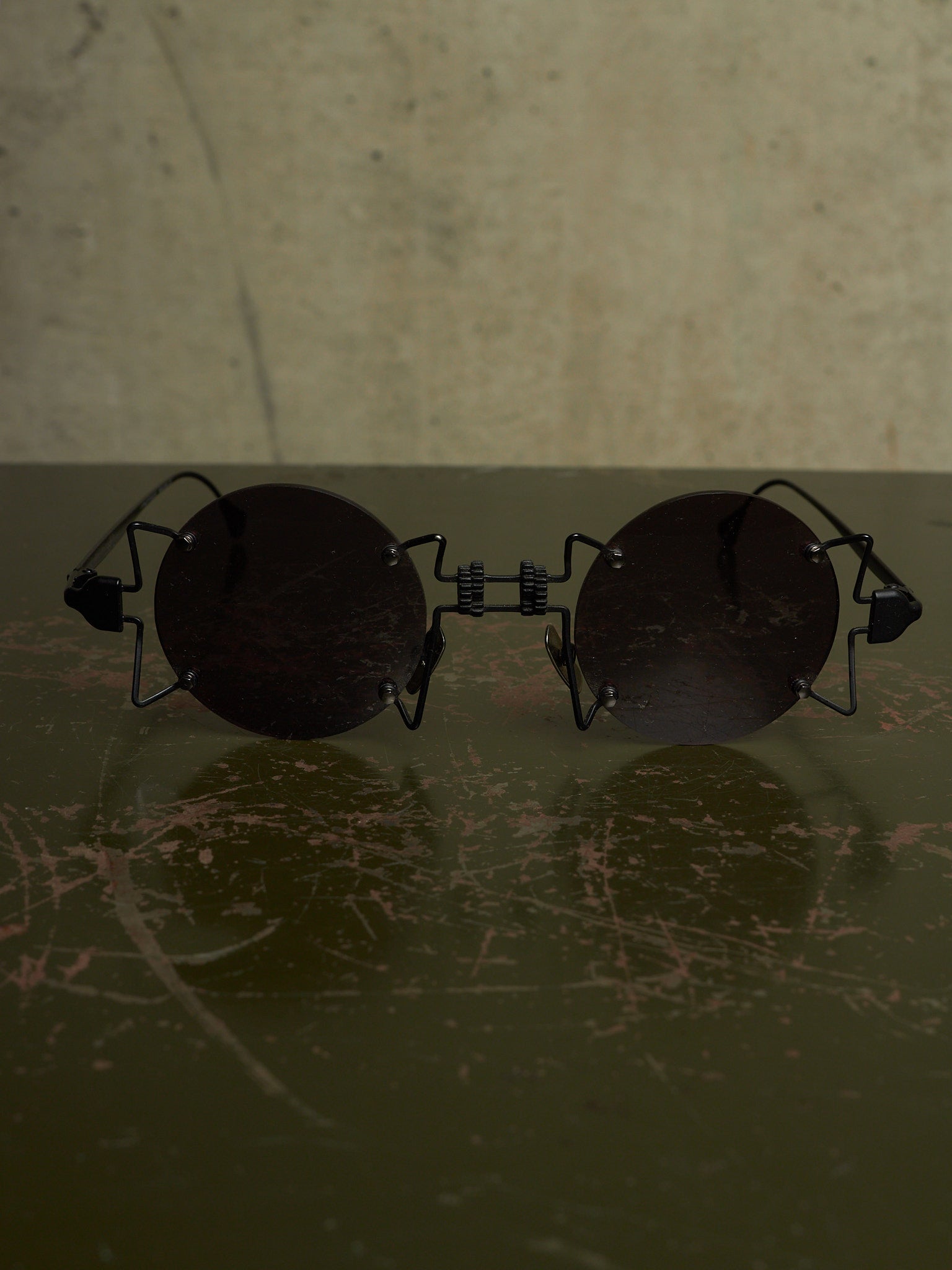 Round black sunglasses with a unique wire frame design, resting on a dark scratched surface against a textured beige background.