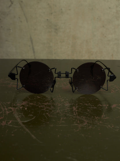 Round black sunglasses with a unique wire frame design, resting on a dark scratched surface against a textured beige background.