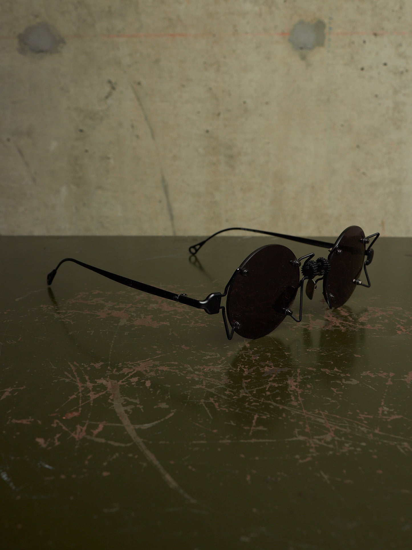 Round black sunglasses with dark lenses resting on a dark reflective surface against a textured concrete wall.