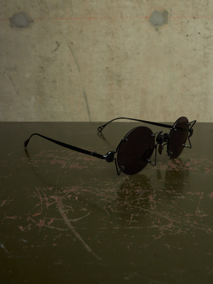 Round black sunglasses with dark lenses resting on a dark reflective surface against a textured concrete wall.