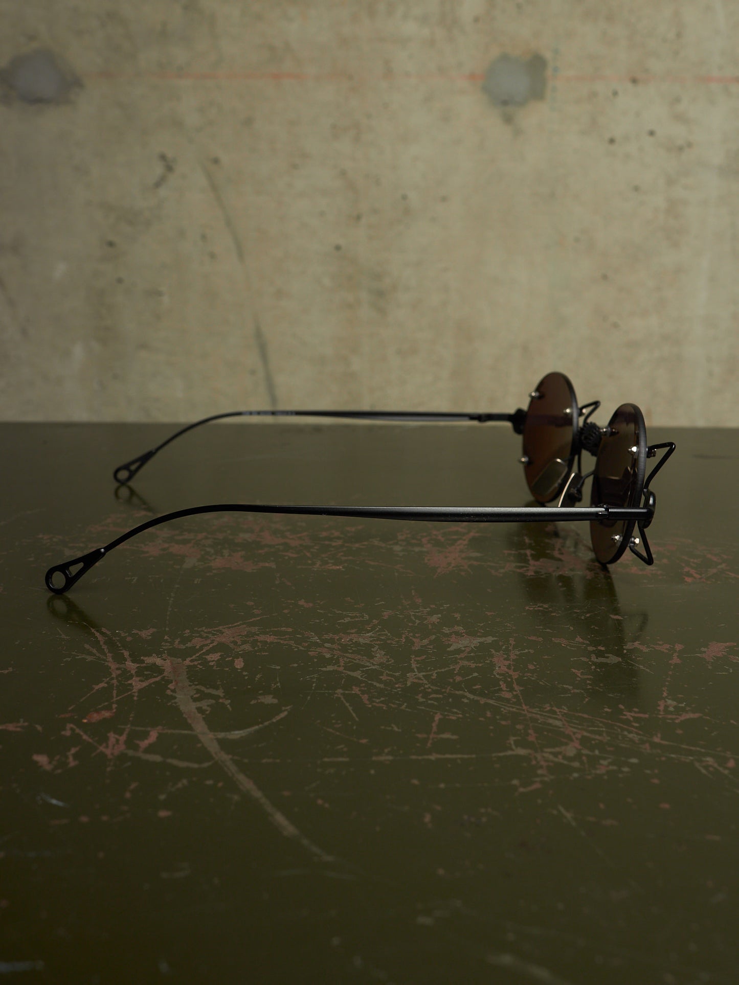 A pair of vintage-style black metal eyeglasses with round brown lenses, resting on a dark, scratched tabletop against a textured concrete wall.