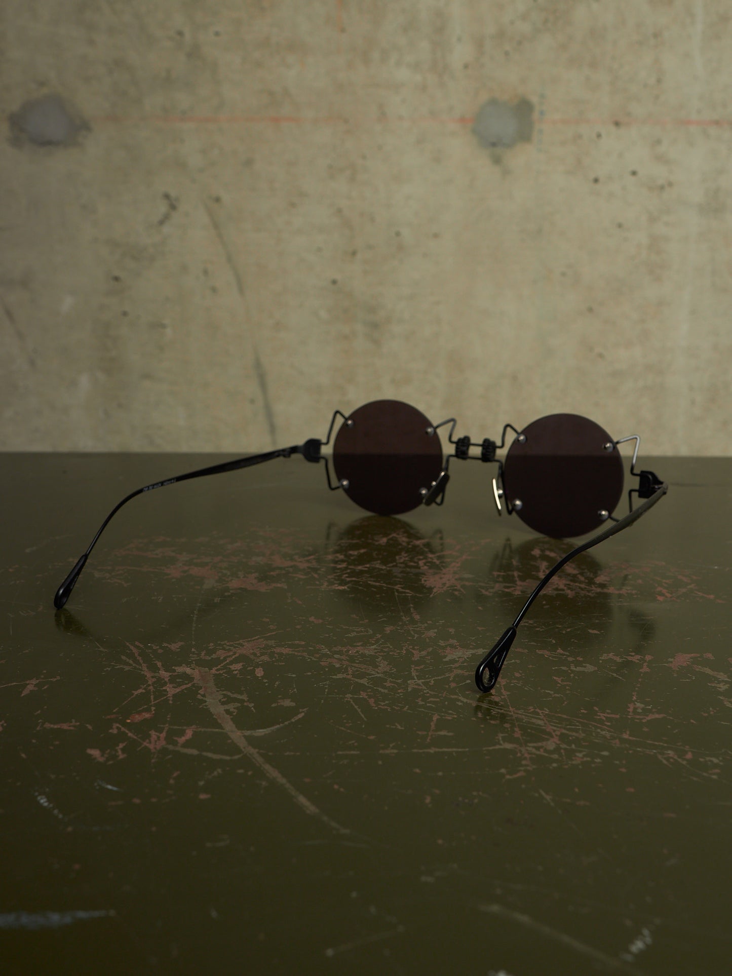 Round black sunglasses with a unique interlocking double-bridge design, resting on a dark scratched surface against a textured concrete wall.