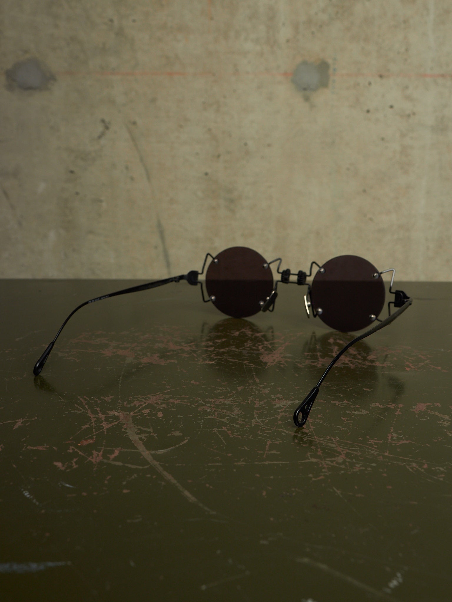 Round black sunglasses with a unique interlocking double-bridge design, resting on a dark scratched surface against a textured concrete wall.