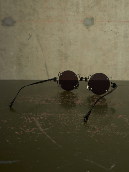 Round black sunglasses with a unique interlocking double-bridge design, resting on a dark scratched surface against a textured concrete wall.