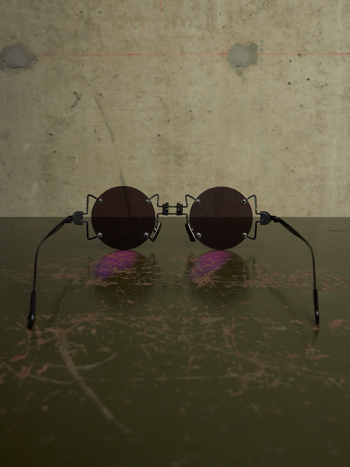 Round sunglasses with dark brown lenses and black metal frames, resting on a dark reflective surface against a textured concrete wall.