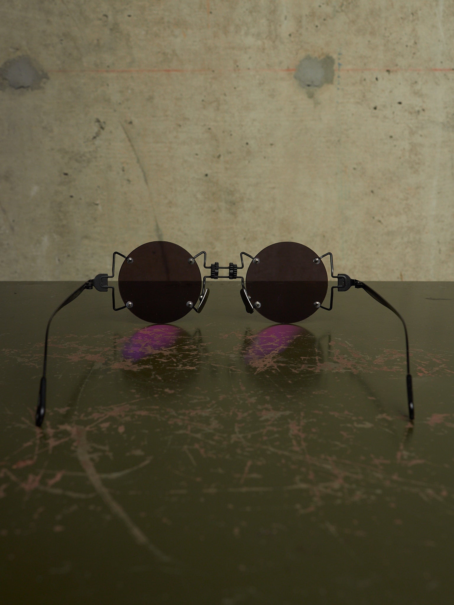Round sunglasses with dark brown lenses and black metal frames, resting on a dark reflective surface against a textured concrete wall.