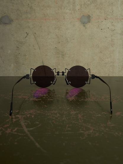 Round sunglasses with dark brown lenses and black metal frames, resting on a dark reflective surface against a textured concrete wall.