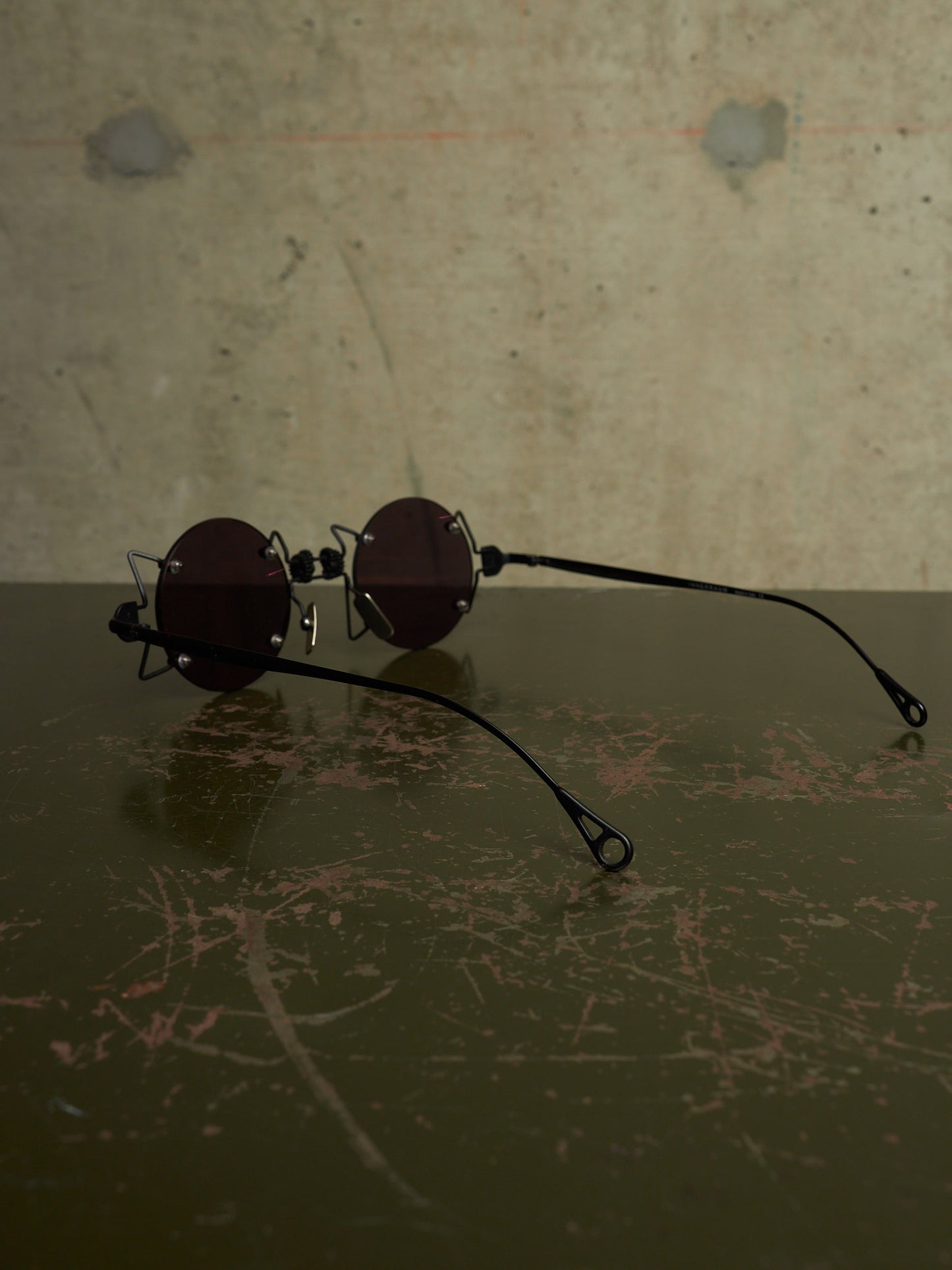 A pair of round dark-lensed sunglasses with thin black metal frames and long black temple arms, resting on a dark scratched surface against a textured concrete wall.
