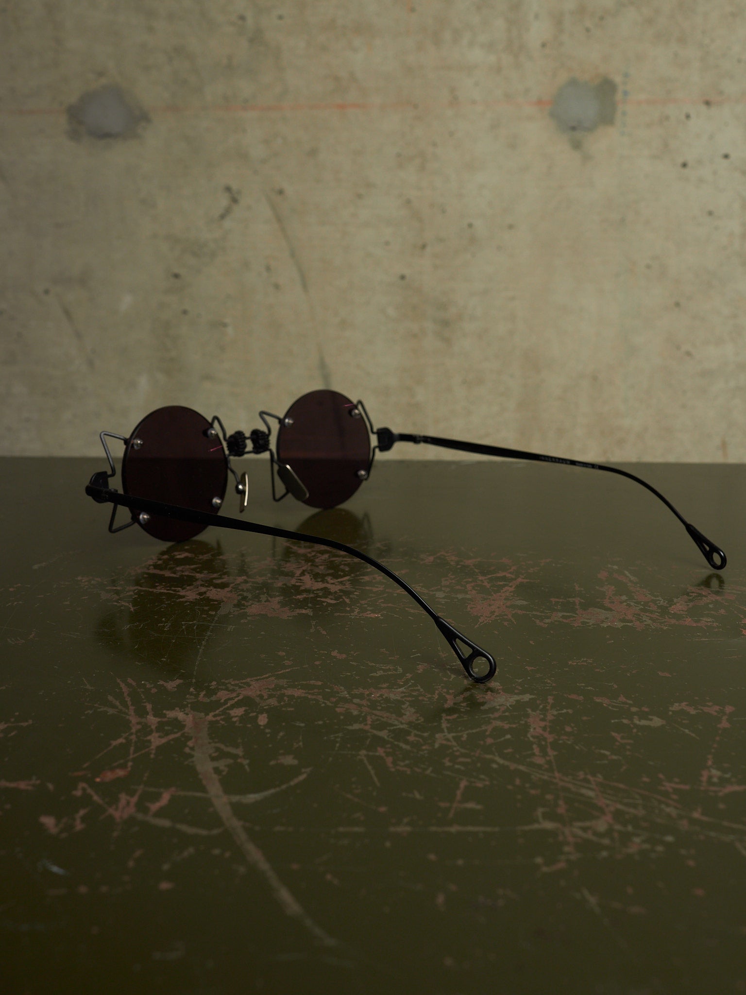 A pair of round dark-lensed sunglasses with thin black metal frames and long black temple arms, resting on a dark scratched surface against a textured concrete wall.