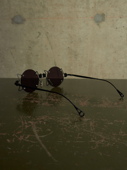 A pair of round dark-lensed sunglasses with thin black metal frames and long black temple arms, resting on a dark scratched surface against a textured concrete wall.