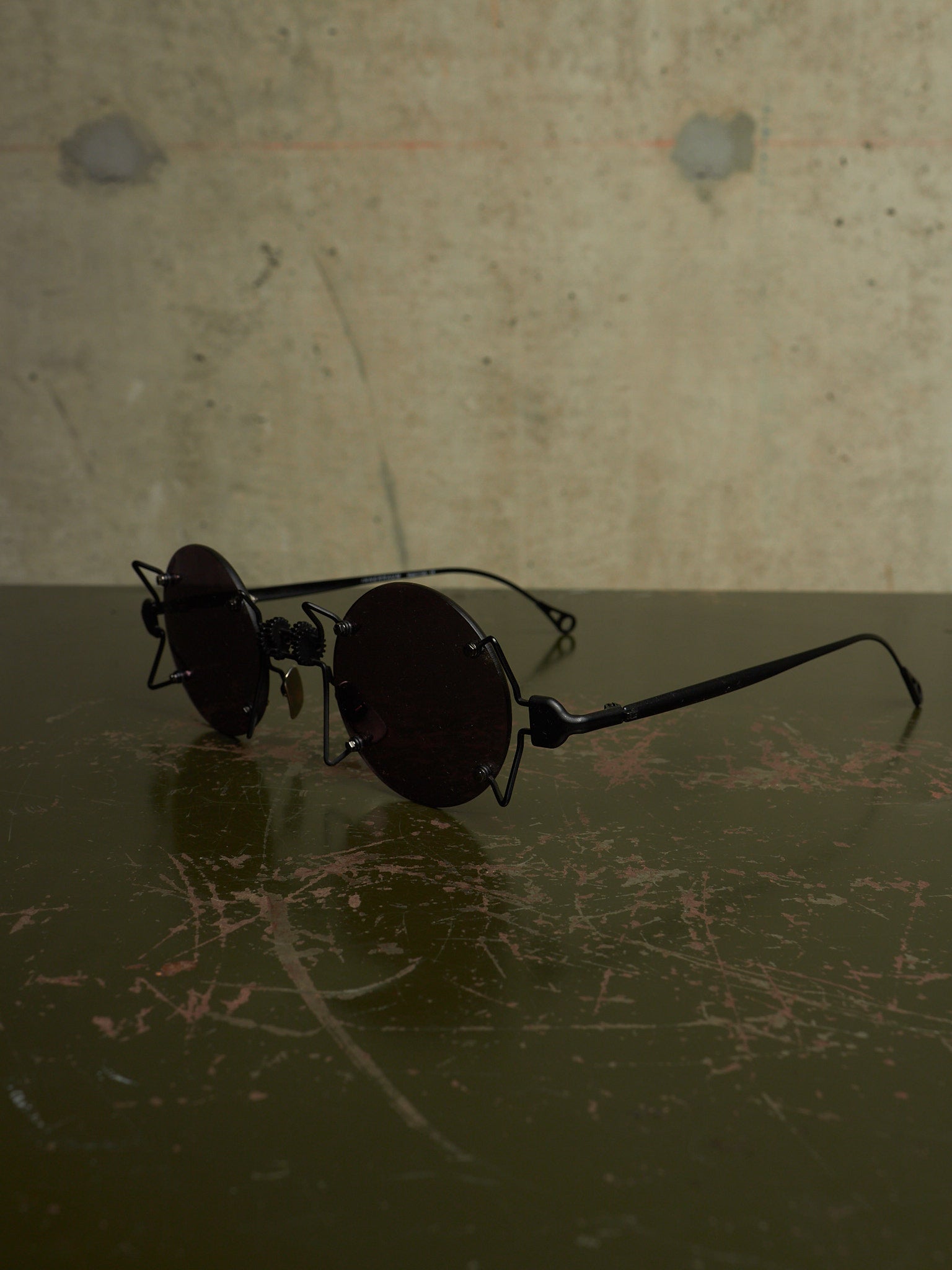 A pair of round black sunglasses with an industrial, vintage aesthetic, featuring a wire frame and dark lenses, resting on a dark reflective surface against a textured concrete wall.