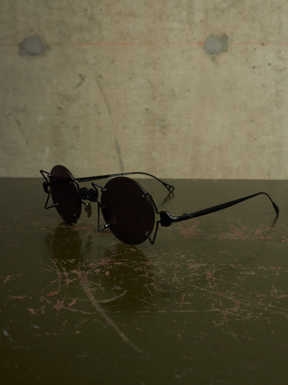 A pair of round black sunglasses with an industrial, vintage aesthetic, featuring a wire frame and dark lenses, resting on a dark reflective surface against a textured concrete wall.