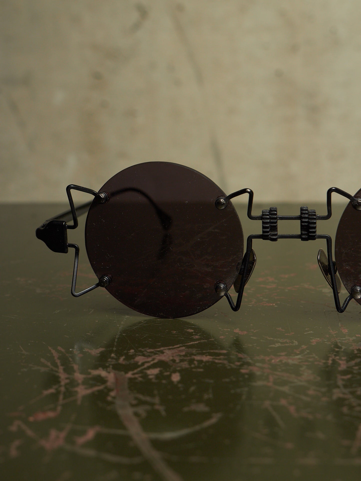 Round dark sunglasses with a unique black metal frame resting on a dark green marble surface against a beige background.