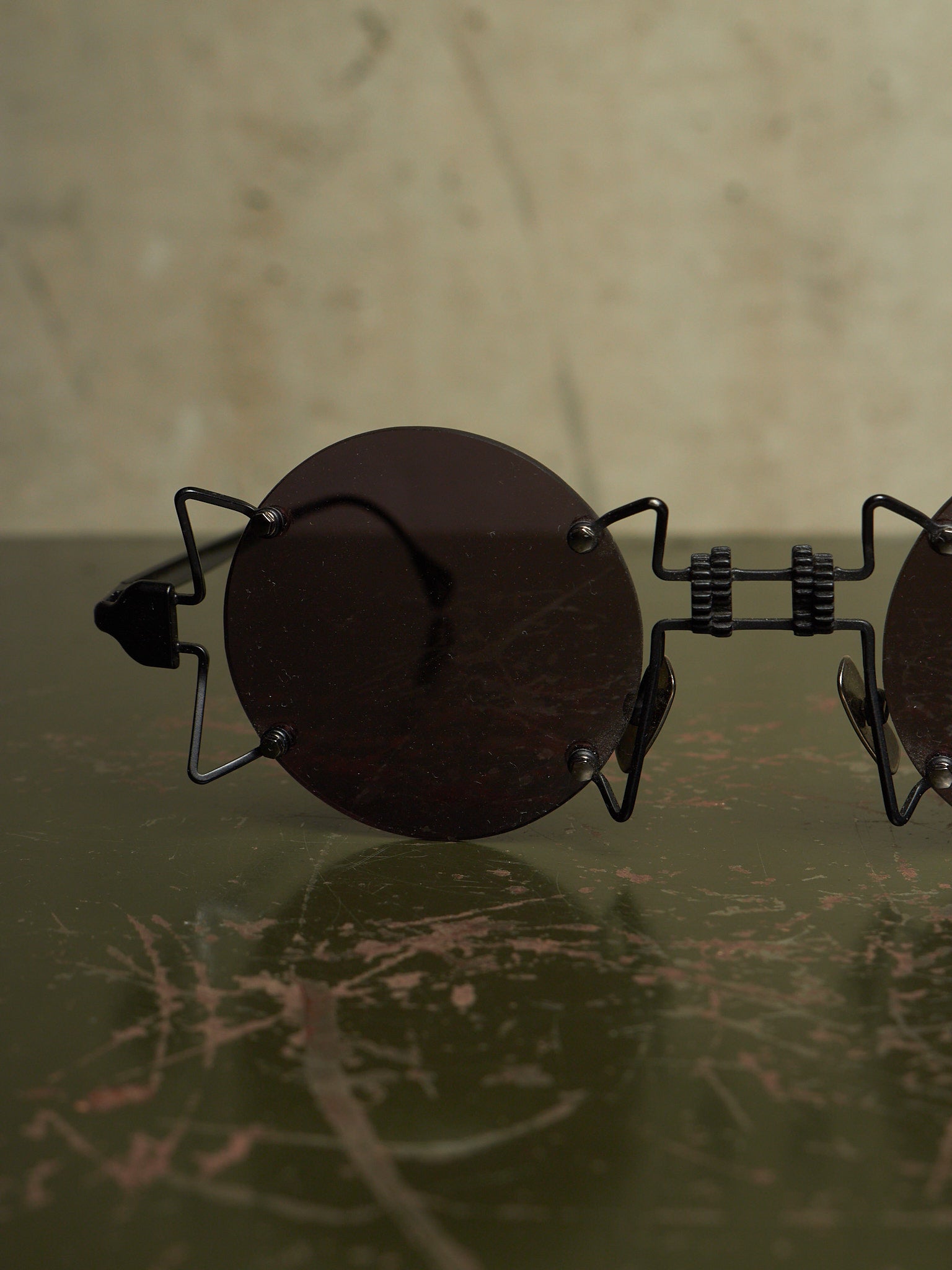 Round dark sunglasses with a unique black metal frame resting on a dark green marble surface against a beige background.