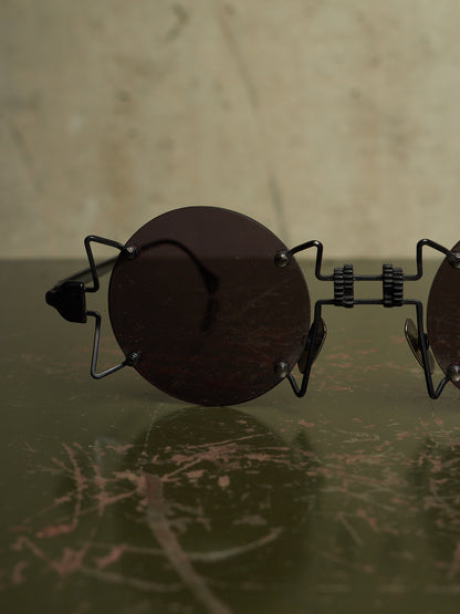 Round dark sunglasses with a unique black metal frame resting on a dark green marble surface against a beige background.