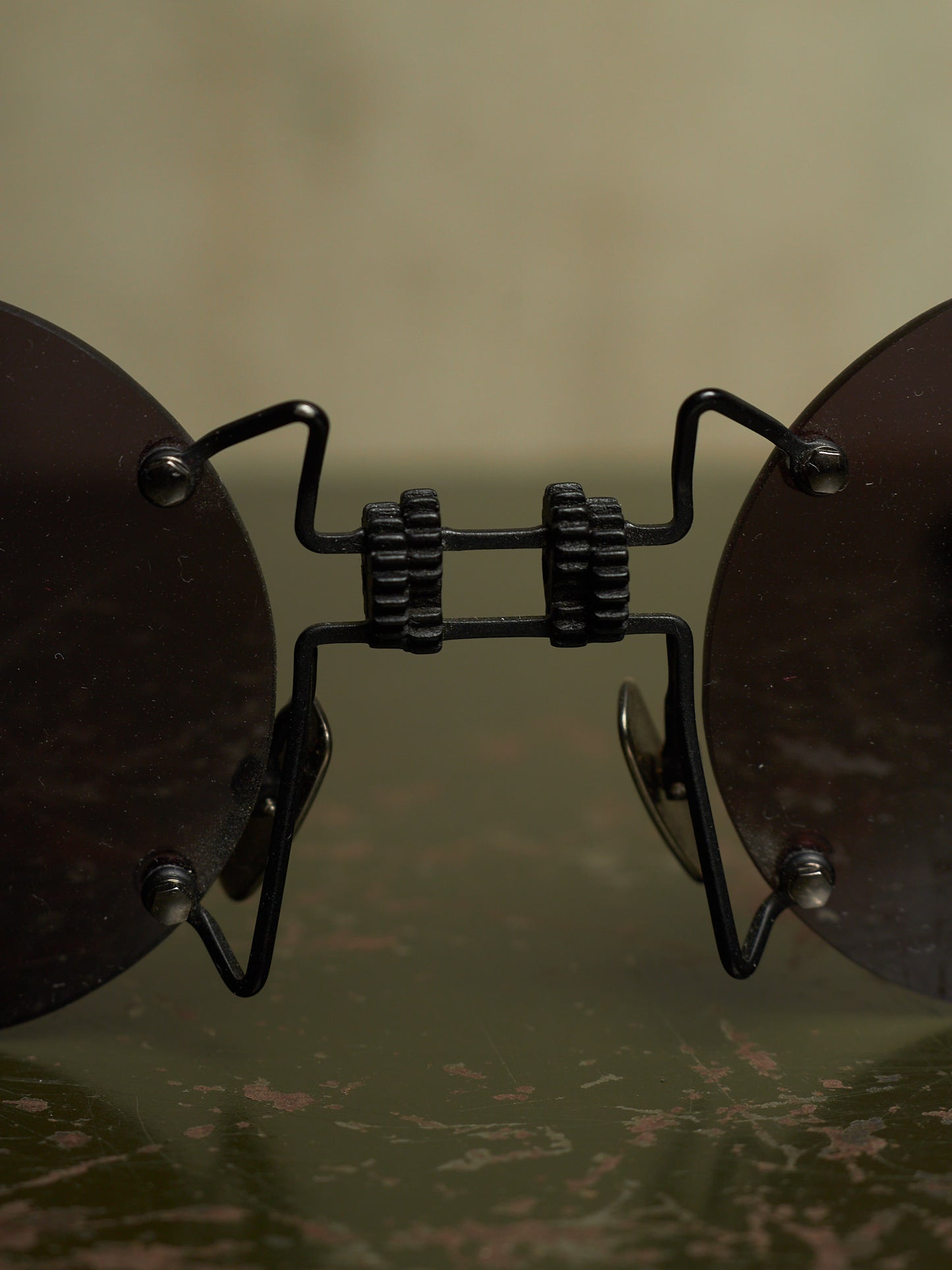 Close-up of a pair of round dark brown sunglasses with a black metal bridge and hinges, resting on a reflective dark surface against a soft beige background.