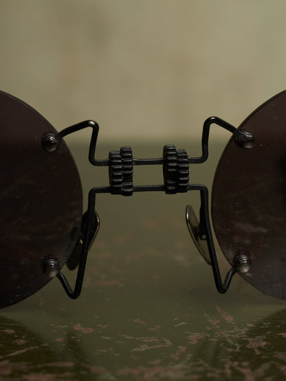 Close-up of a pair of round dark brown sunglasses with a black metal bridge and hinges, resting on a reflective dark surface against a soft beige background.