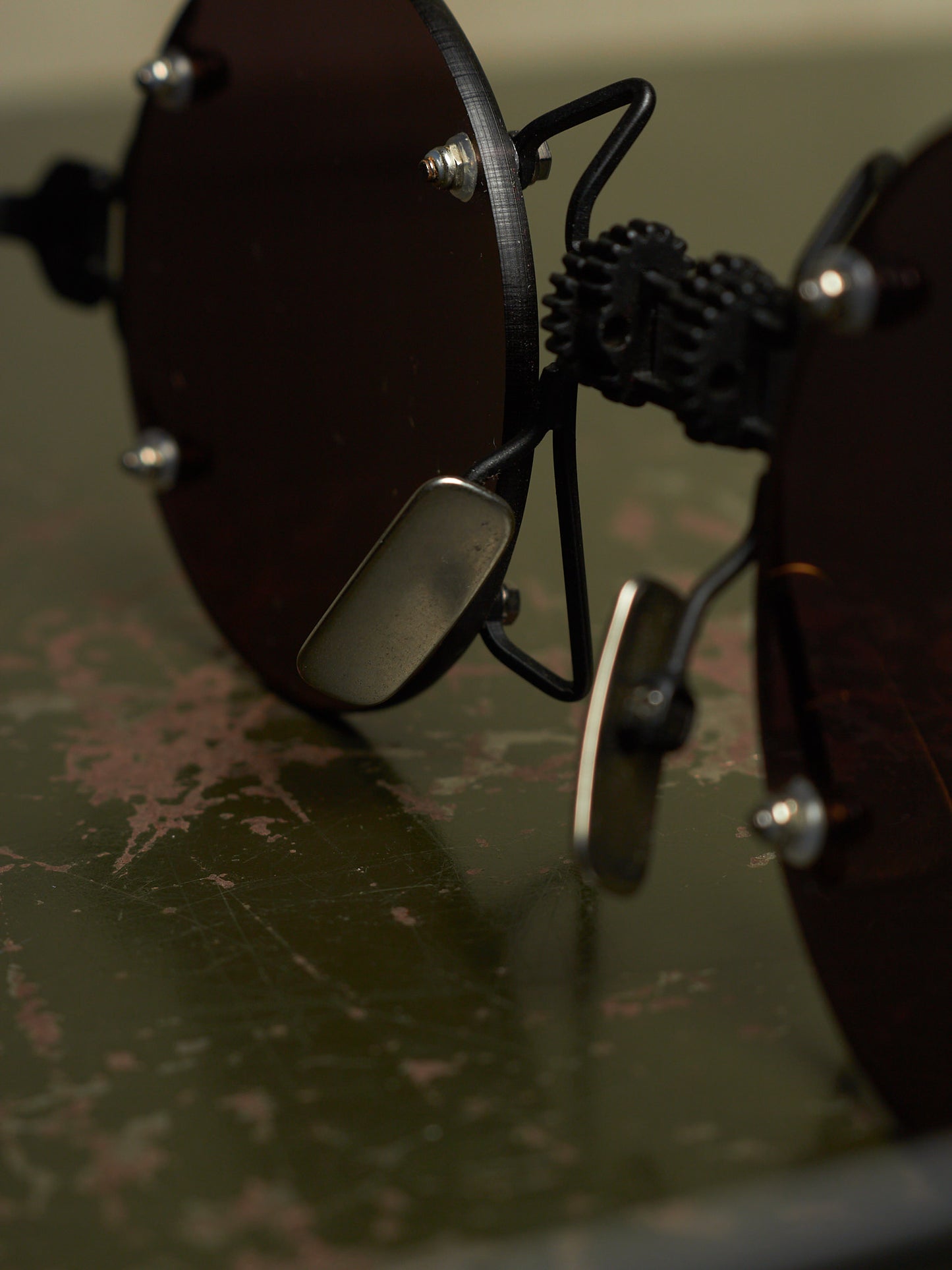 Close-up of a pair of dark tinted rimless sunglasses with a unique black mechanical frame and small silver metal nose pads, resting on a distressed green surface.