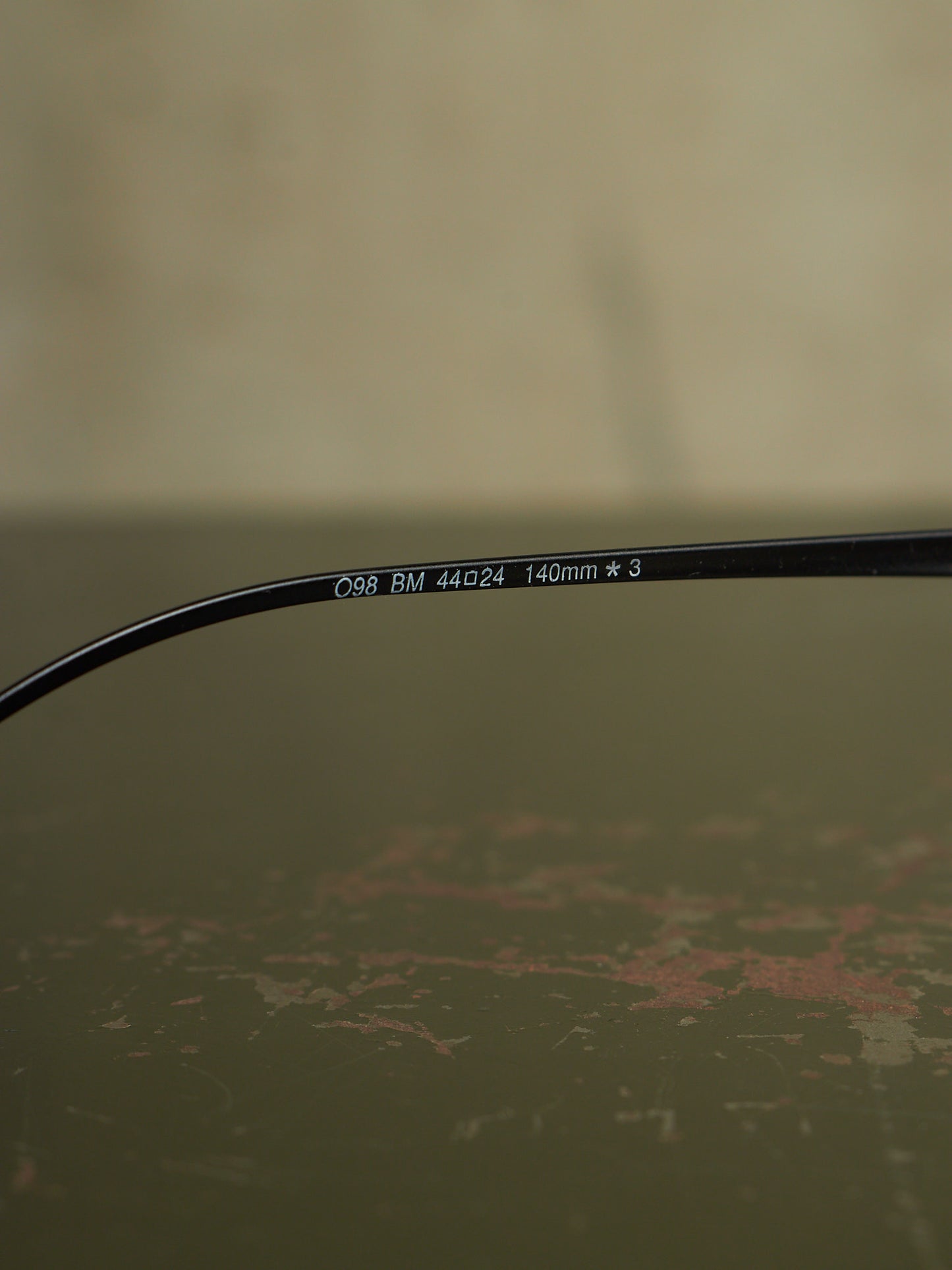 Close-up of a black eyeglass temple arm with white printed text reading 'C98 BM 44a24 140mm * 3' on a textured olive-green surface.