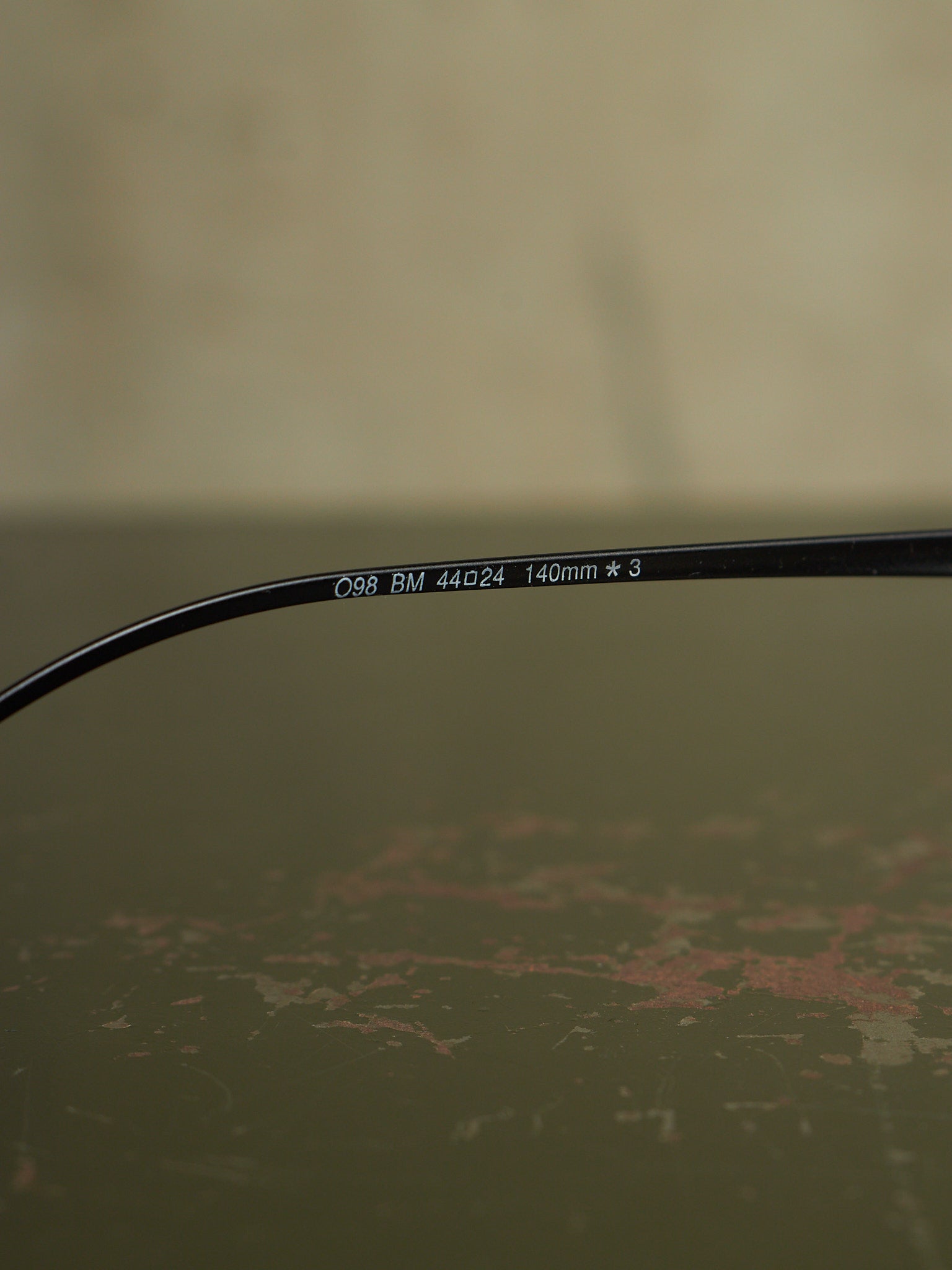 Close-up of a black eyeglass temple arm with white printed text reading 'C98 BM 44a24 140mm * 3' on a textured olive-green surface.