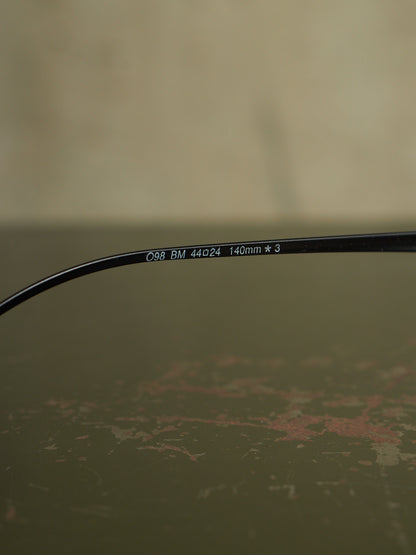 Close-up of a black eyeglass temple arm with white printed text reading 'C98 BM 44a24 140mm * 3' on a textured olive-green surface.