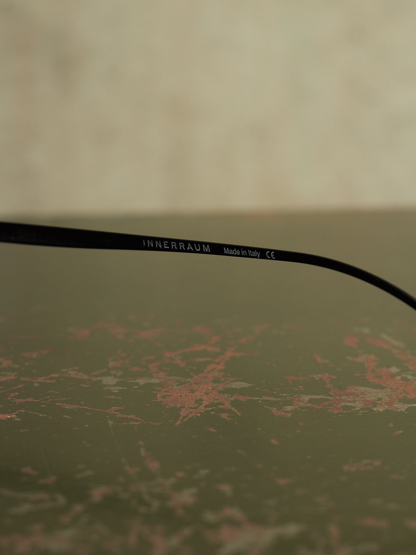 A close-up of a black optical temple arm with the brand name 'INNERRAUM' printed in white, resting on a textured, distressed olive-green surface.