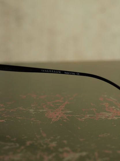 A close-up of a black optical temple arm with the brand name 'INNERRAUM' printed in white, resting on a textured, distressed olive-green surface.