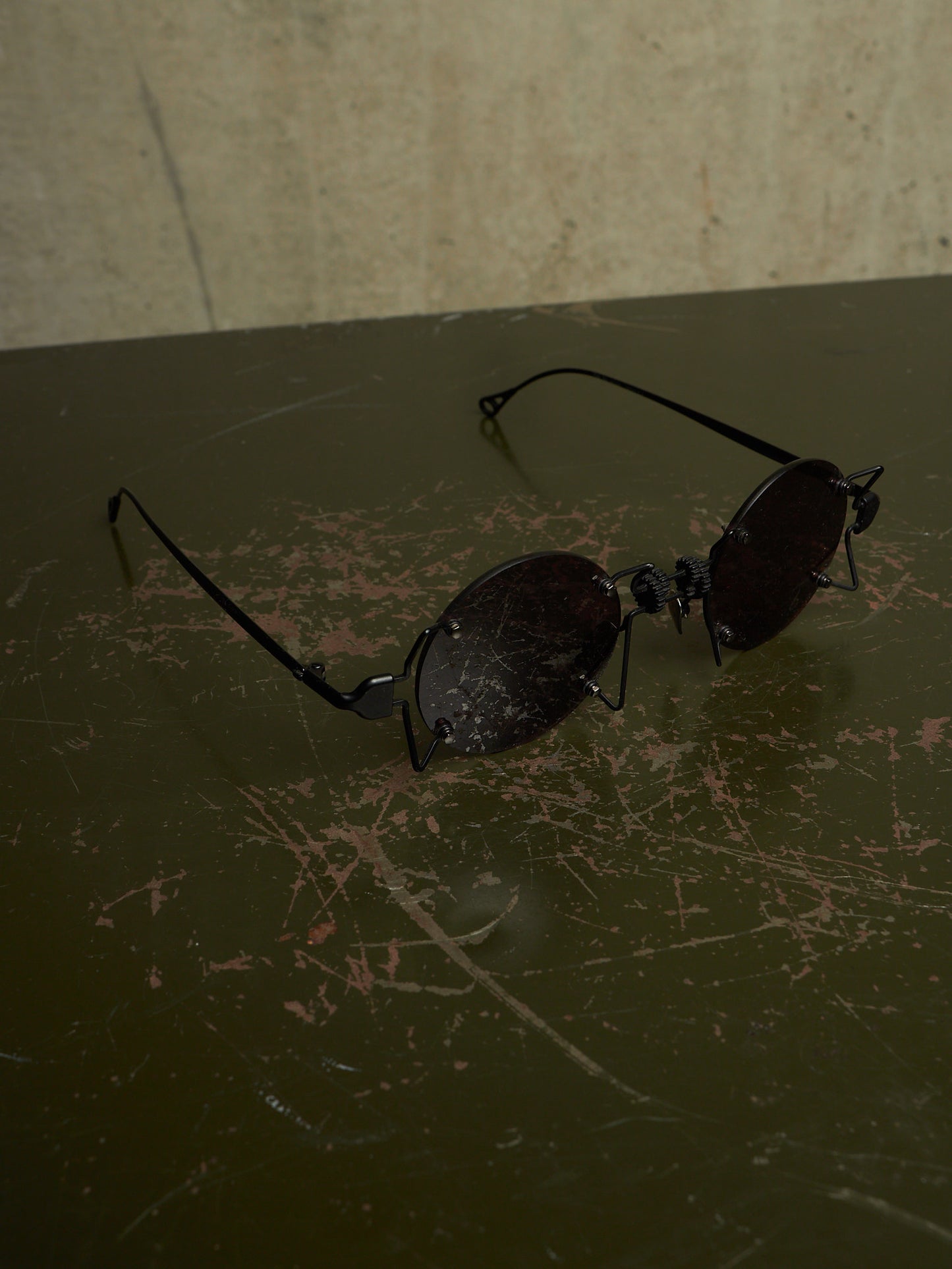 A pair of black metal-framed sunglasses with dark tinted lenses resting on a dark, scratched tabletop against a textured beige wall.