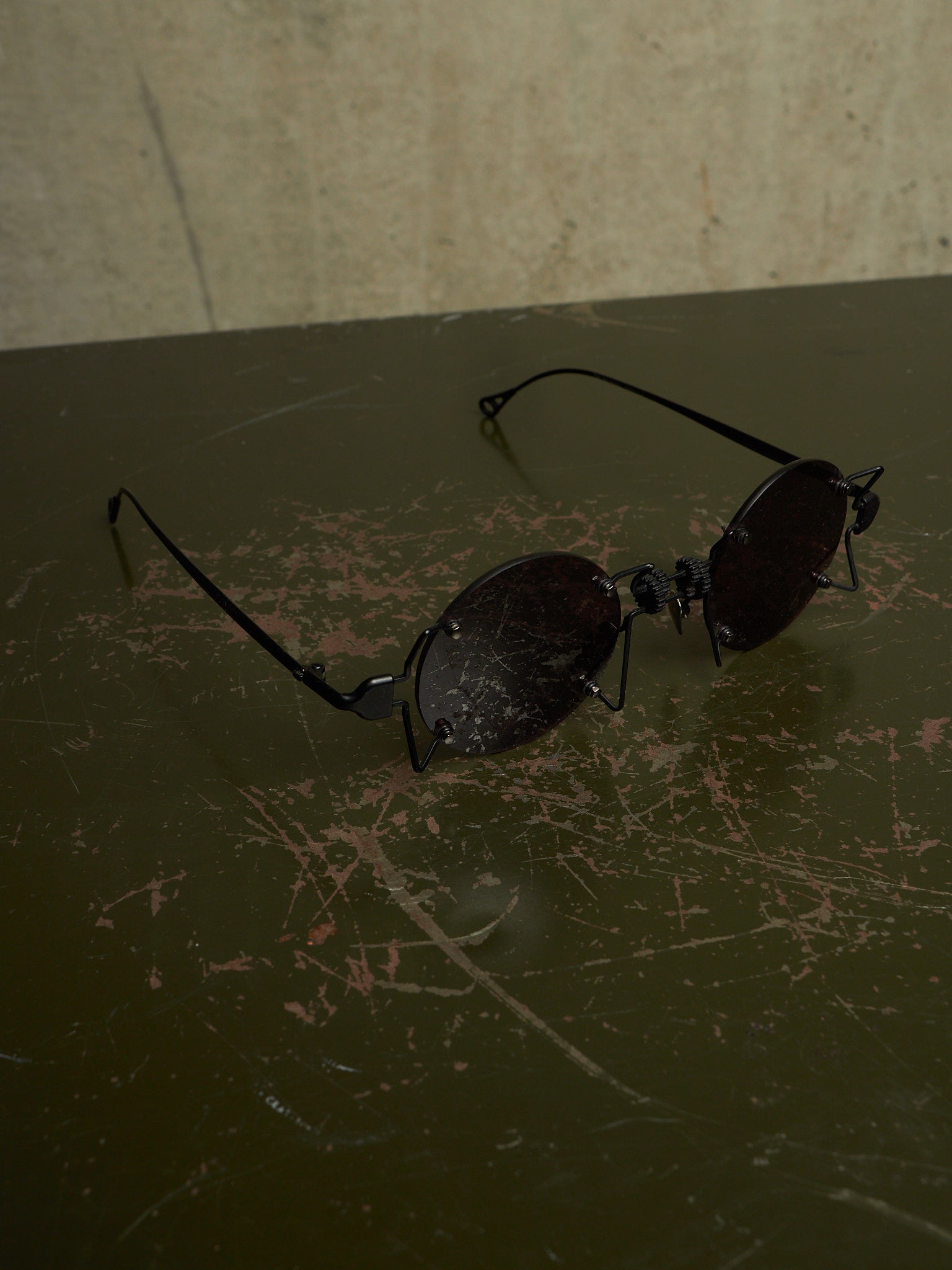 A pair of black metal-framed sunglasses with dark tinted lenses resting on a dark, scratched tabletop against a textured beige wall.