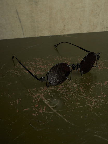 A pair of black metal-framed sunglasses with dark tinted lenses resting on a dark, scratched tabletop against a textured beige wall.