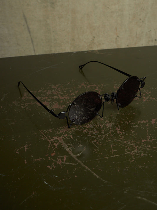 A pair of black metal-framed sunglasses with dark tinted lenses resting on a dark, scratched tabletop against a textured beige wall.